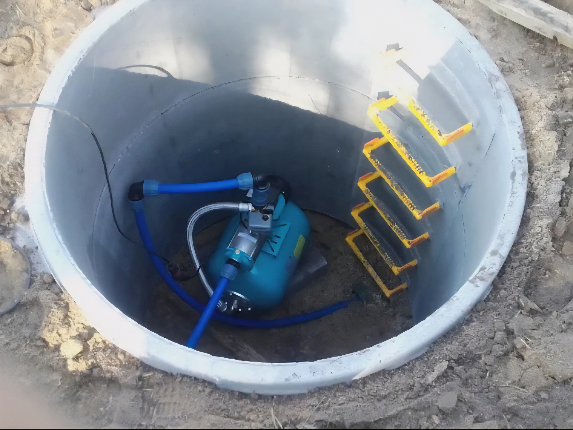 Wnętrze studni z betonowego kręgu z niebieskim hydroforem, wężami i drabiną